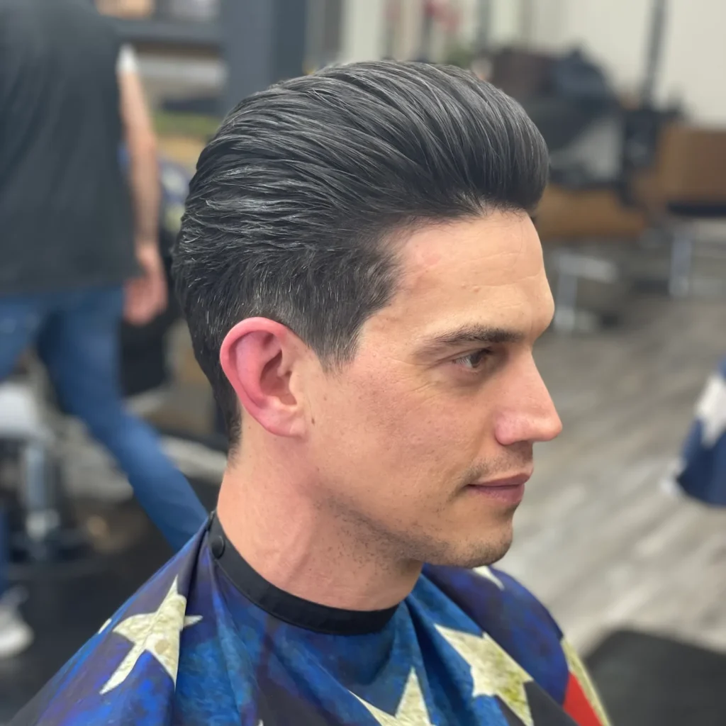 Man in a barber chair with a slicked-back pompadour and tapered sides, seen from the side